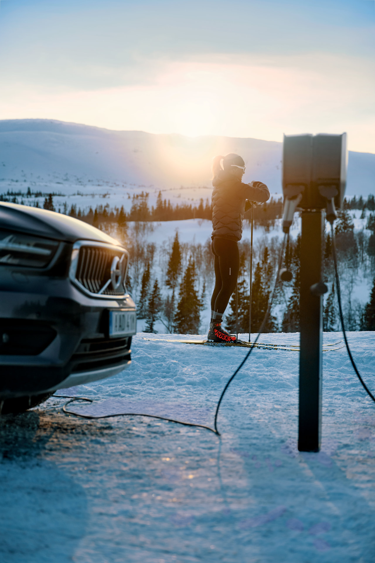 En elbil ansluten till en laddstation bredvid en skidåkare i ett snötäckt fjällandskap vid soluppgången.